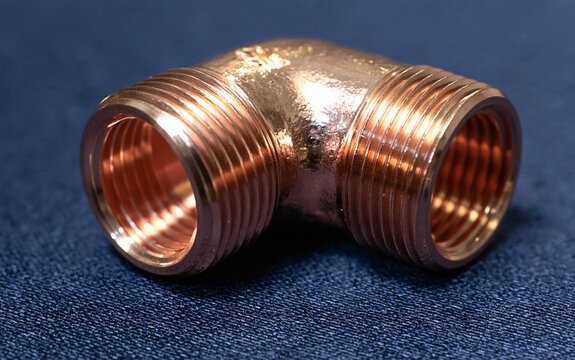 Polished copper elbow pipe fitting with internal threads rests on a dark blue surface. This plumbing component connects pipes at a right angle for water systems.