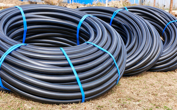 Coiled black polyethylene pipes for water supply, irrigation systems sit on ground. Bundled with blue plastic straps, ready for construction project use.