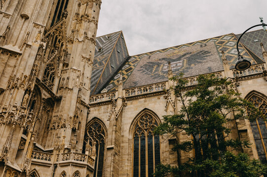 ornate gothic cathedral exterior under overcast sky, patterned tiled roof, tall arched stained glass windows.