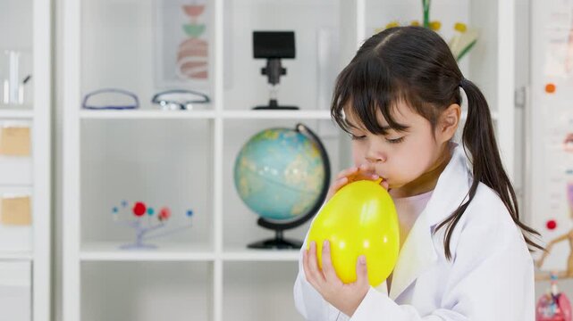 Asian Schoolgirl In Lab Coat Blowing Up Yellow Balloon In Science Classroom