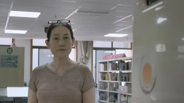An adult woman stands in a well-lit library interior with bookshelves, windows, and ceiling fixtures.