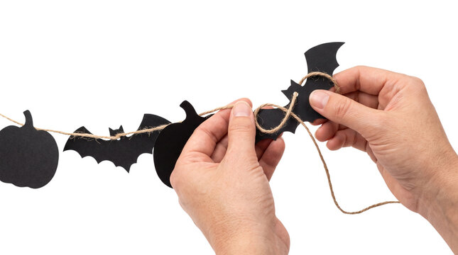 Halloween DIY crafts, human hands stringing black paper bats and pumpkin garland on twine string, handmade party decoration concept