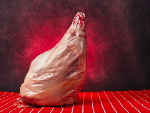 Raw lamb leg in plastic vacuum wrap rests on red and white table cloth, painted background. Meat shows pink flesh and red fat marbling. Scene suggests butchery, cooking, or grocery retail.