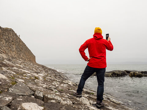 Man in red jacket stands alone on rocky coast, facing misty sea. Yellow beanie adds pop of color against gray sky. He appears contemplative, gazing at water and distant horizon