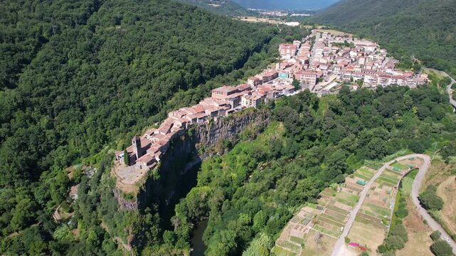 Forward reveal aerial shot of Castellfollit de la Roca, a stunning medieval village perched on a narrow basalt cliff crag between two rivers. Girona, Catalonia, Spain.
