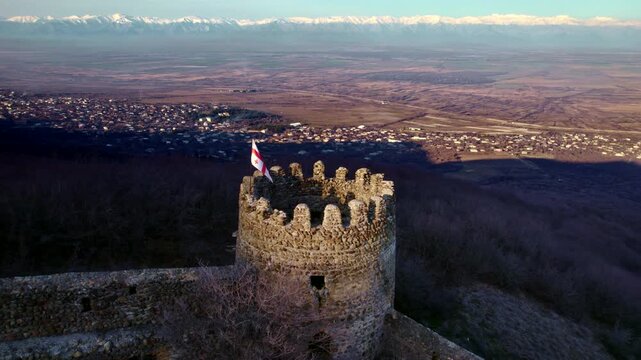 Round fortress tower and Georgian flag dominate the foreground as the drone shifts to reveal more of the Alazani plain with scattered settlements brown fields and a long snowy mountain range