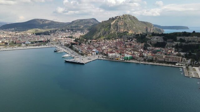 Nafplio coastal town with historic fortress on rocky hill overlooking harbor and bay. Aerial view captures waterfront promenade, marina, and red-roofed buildings against mountain backdrop.