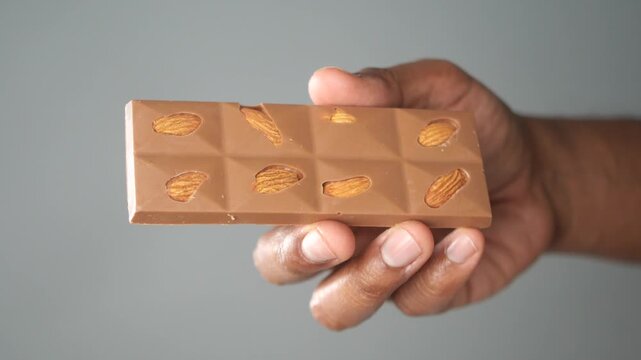 Close up of hand holding broken chocolate bar with almonds against gray background.