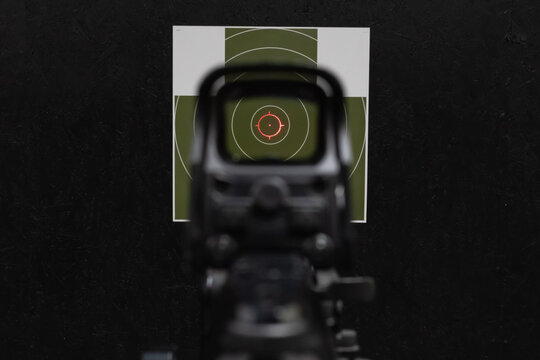 First person view through holographic sight aiming red dot reticle at paper target on black bullet trap in indoor shooting range