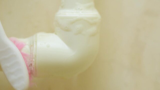 Closeup plastic pipe corner cleaned with toothbrush. Pink bristles scrub white PVC elbow at high speed, producing foam and debris removal. Close framing, shallow depth of field,
