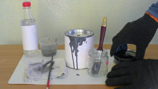 Right gloved hand pours clear solvent from plastic bottle into small jar next to upright paint brush and open paint can with dark gray drips on white exterior, with paint, brush, solvent, jar detail