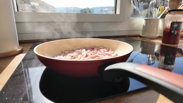 Close view of diced bacon steaming in a frying pan on a sunlit induction cooktop beside a kitchen window.