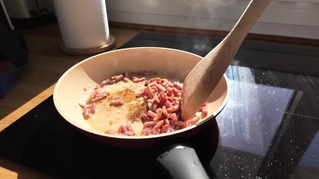Close-up of diced bacon frying in a cream-colored pan with a wooden spoon on an induction cooktop.