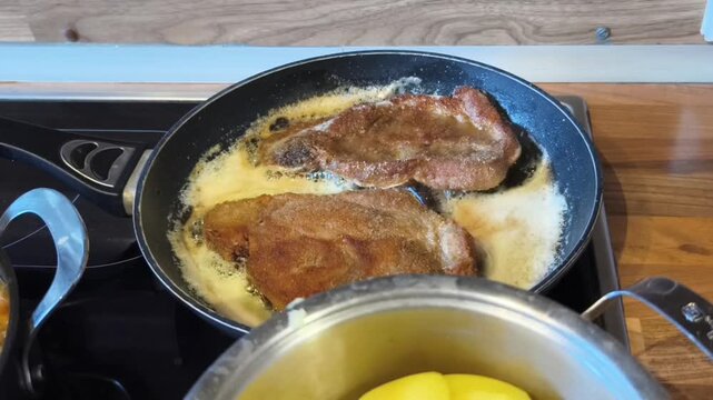 2 panierte Koteletts braten goldbraun in Fett in einer Bratpfanne auf dem Herd. Nahaufnahme, Echtzeit, Originalton. Vorne Kartoffeln und Linsengem&uuml;se. 