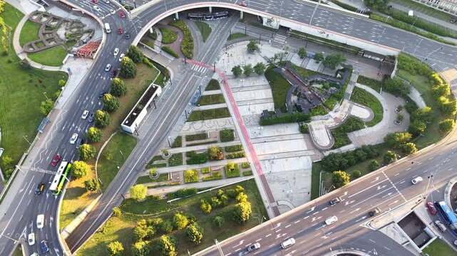 Aerial view of a busy urban transport interchange showcasing vehicles navigating multiple roadways, with green spaces and pedestrian pathways visible in a bustling city environment. Modern city