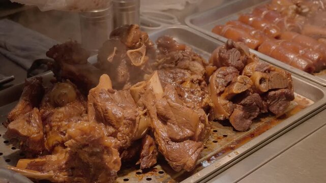 Cooked bone-in meat and braised meat displayed on perforated metal trays in a food service setting.