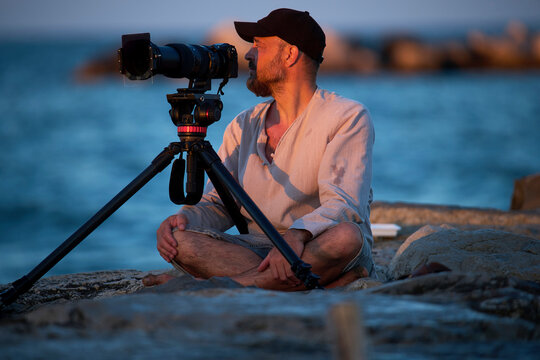 Man sits near ocean with camera and tripod