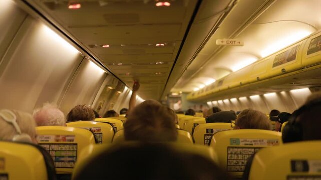 Interior of a commercial airplane during flight with travelers sitting in multiple rows of seats surrounded by warm ambient lighting inside the passenger cabin