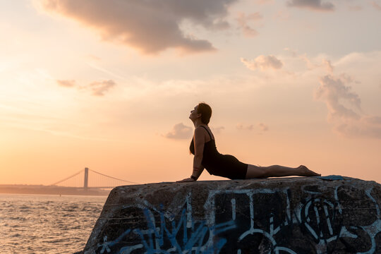Woman holding a prone backbend on a concrete platform. A woman presses both palms down and lifts her head beneath orange clouds above water and a bridge.