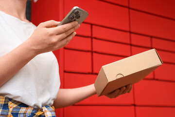 Naklejka na ściany i meble Parcel delivery and logistics, woman using smartphone to collect package from automated red locker terminal for online shopping and e-commerce convenience