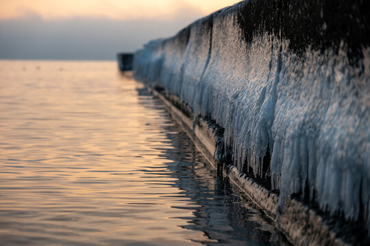 frozen pier covered with ice and icicles at sea sunset, icy concrete breakwater on calm ocean water during winter sunrise, close up view of ice covered pier wall with golden water reflections