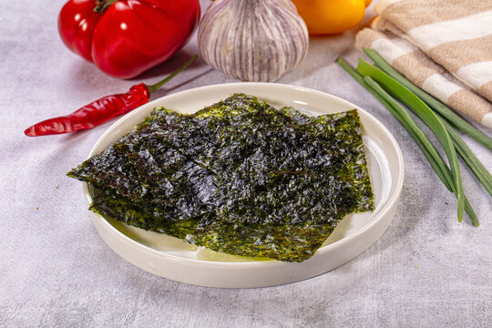 Ceramic plate holding dark green nori sheets surrounded by fresh vegetables on grey surface Asian cuisine ingredient
