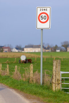 A European "ZONE 50" speed limit traffic sign stands next to a rural road, overlooking green grassy fields, a fence, and distant village houses under a clear sky.