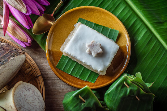 Traditional Thai tako taro pudding dessert square cut topped with thick coconut milk cream in aluminum tray next to banana leaf parcels and fresh sliced taro root. Asian sweet food snack.
