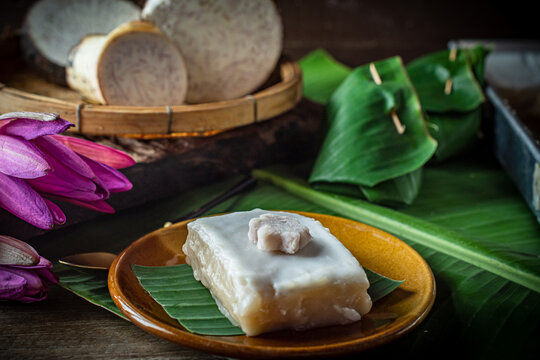 Traditional Thai tako taro pudding dessert square cut topped with thick coconut milk cream in aluminum tray next to banana leaf parcels and fresh sliced taro root. Asian sweet food snack.