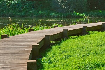 Naklejka premium Winding wooden boardwalk path along a peaceful pond in a sunny park. Scenic rural landscape with fresh green grass and natural timber pedestrian walkway on a summer day.