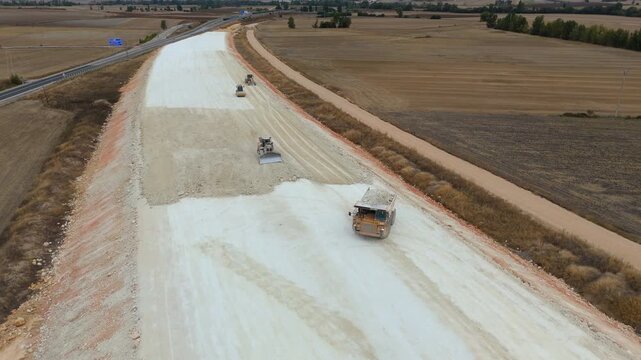 An aerial view of heavy machinery including a dump truck unloading soil, a bulldozer, and a grader working on a road construction embankment