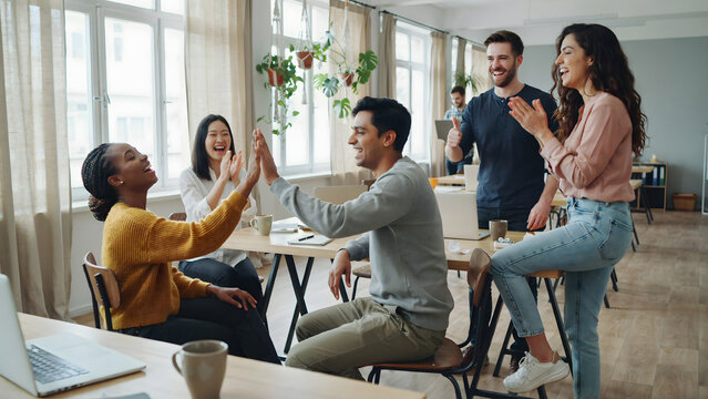 A diverse group of young colleagues in a bright office laughing and giving fives to celebrate a successful team project and collaboration