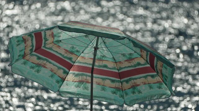 Palm patterned parasol ripples under a steady breeze above sparkling ocean waves during a bright summer day. Static medium shot records the scene.