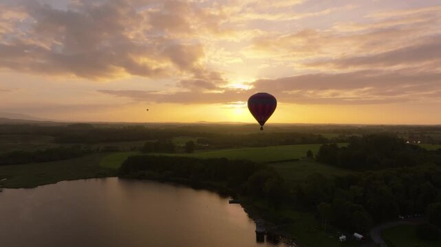 A colorful air balloon drifts through a glowing sunset sky above calm lakeside farmland and dark tree lines, filmed from a drone.