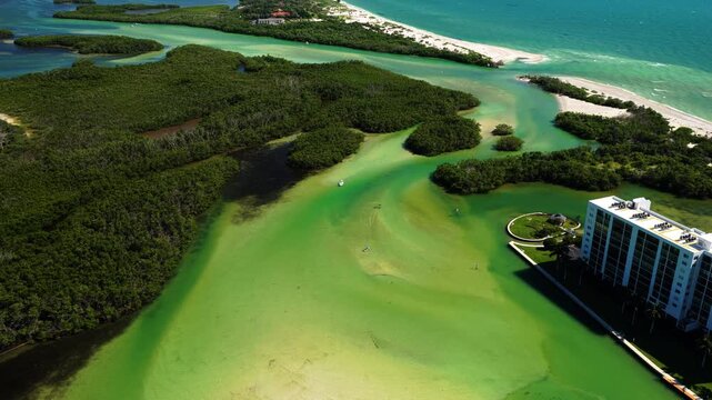 Mangrove islands and winding channels border shallow green water, with a sandy ocean beach and a multistory waterfront building nearby.