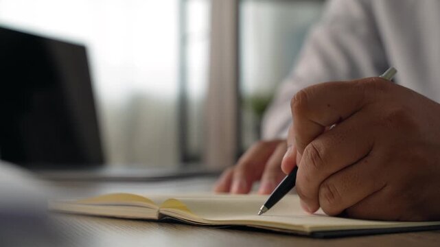 Hands writing notes in a notebook beside a laptop, showing business planning, record keeping and budget preparation at an office desk.