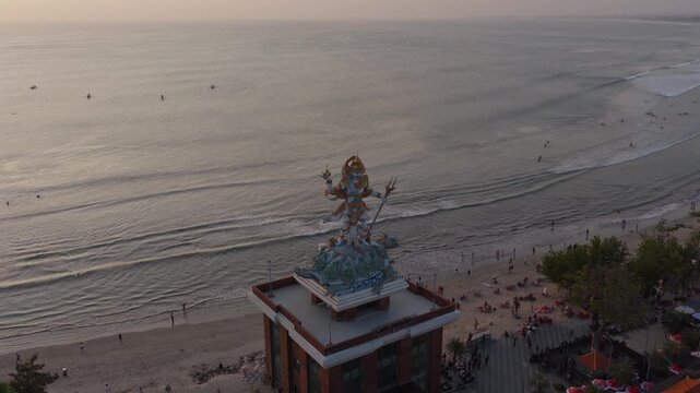 Dewa Baruna Hindu god statue overlooking Kuta beach coastline and rolling ocean surf at sunset in Bali iconic Indonesian travel landmark push-in aerial drone view