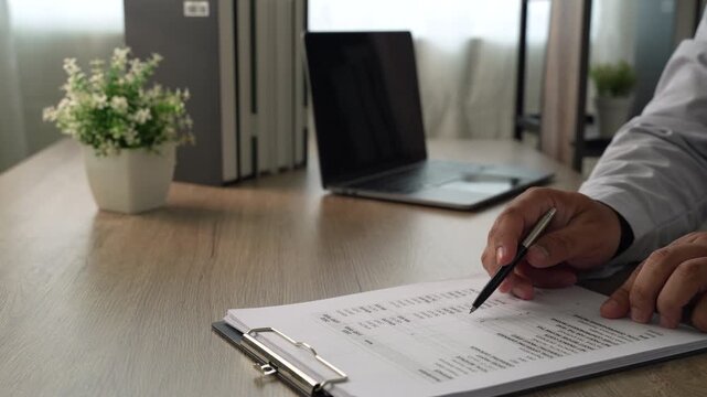 Hands reviewing financial tables on a clipboard with pen beside a laptop, showing accounting analysis, budget review and financial data checking.