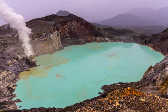 Ijen volcano complex in East Java, Indonesia, with a turquoise acidic crater lake, rugged rock walls, yellow sulfur deposits, and thick white smoke rising from active vents.
