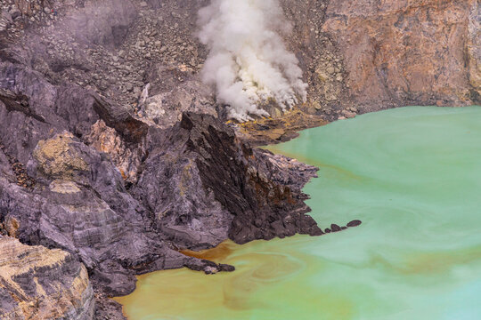 Ijen volcano complex in East Java, Indonesia, with a turquoise acidic crater lake, rugged rock walls, yellow sulfur deposits, and thick white smoke rising from active vents.