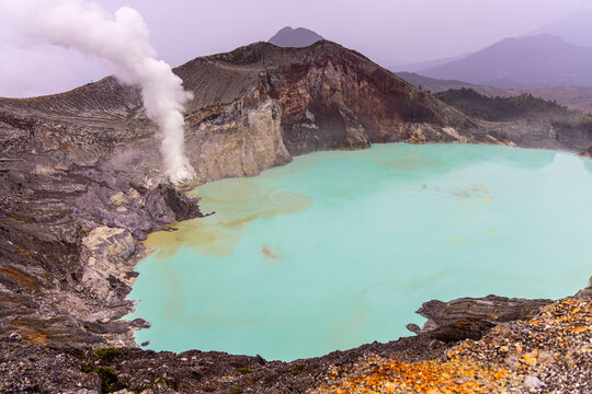 Ijen volcano complex in East Java, Indonesia, with a turquoise acidic crater lake, rugged rock walls, yellow sulfur deposits, and thick white smoke rising from active vents.