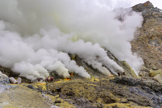 Ijen volcano complex in East Java, Indonesia, with a turquoise acidic crater lake, rugged rock walls, yellow sulfur deposits, and thick white smoke rising from active vents.