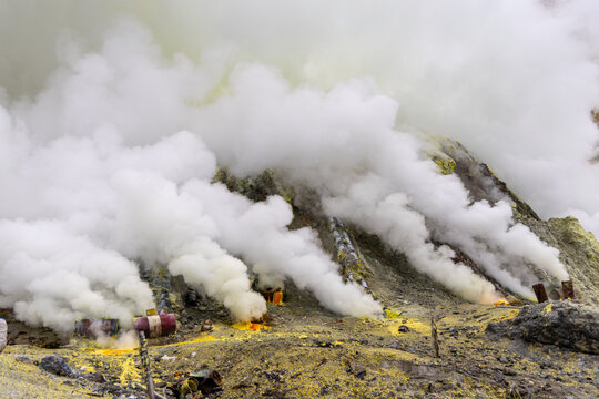 Ijen volcano complex in East Java, Indonesia, with a turquoise acidic crater lake, rugged rock walls, yellow sulfur deposits, and thick white smoke rising from active vents.
