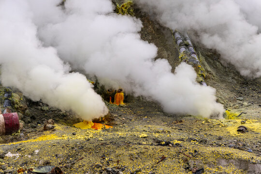 Ijen volcano complex in East Java, Indonesia, with a turquoise acidic crater lake, rugged rock walls, yellow sulfur deposits, and thick white smoke rising from active vents.