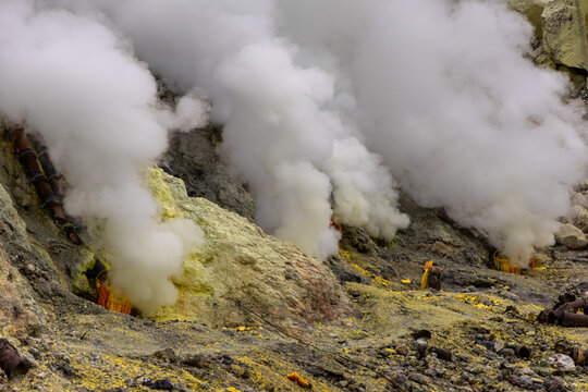 Ijen volcano complex in East Java, Indonesia, with a turquoise acidic crater lake, rugged rock walls, yellow sulfur deposits, and thick white smoke rising from active vents.