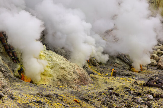 Ijen volcano complex in East Java, Indonesia, with a turquoise acidic crater lake, rugged rock walls, yellow sulfur deposits, and thick white smoke rising from active vents.
