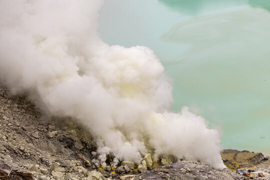 Ijen volcano complex in East Java, Indonesia, with a turquoise acidic crater lake, rugged rock walls, yellow sulfur deposits, and thick white smoke rising from active vents.