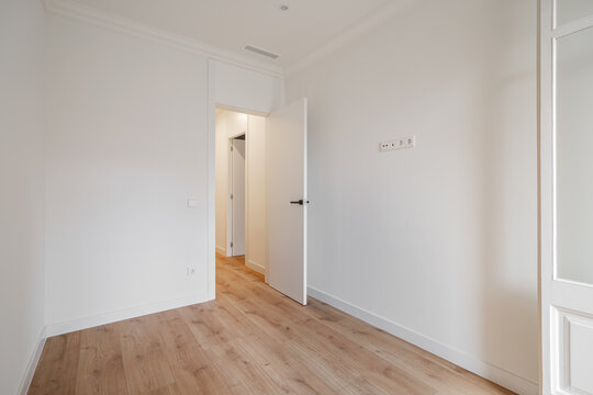 Empty room with white walls and wooden floor. A door is slightly open, leading to another space. Natural light illuminates the area.