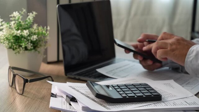 Hands using calculator and pen with printed financial documents beside laptop, showing accounting calculation, financial data analysis and report review.