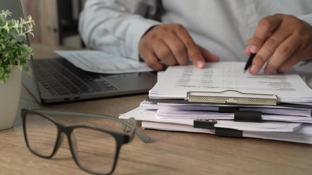 Hands pointing at financial tables on clipboard beside laptop and stacked documents, showing accounting analysis, budget review and financial report checking.